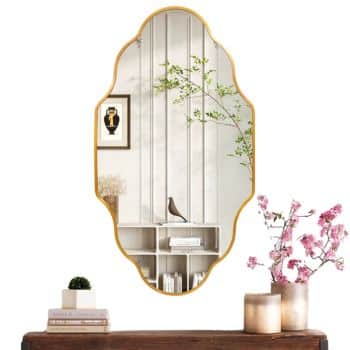 A decorative mirror with a gold frame reflects a minimalist room with a potted plant, a small sculpture, and an art print on the wall. On the wooden table in front of the mirror, there are pink flowers and candles.