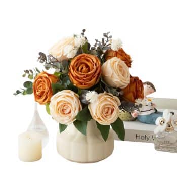 A floral arrangement of peach and rust-colored roses in a white vase, accompanied by greenery and small white blooms. Nearby, there's a lit white candle, a decorative figurine, and a book titled "Holy W.