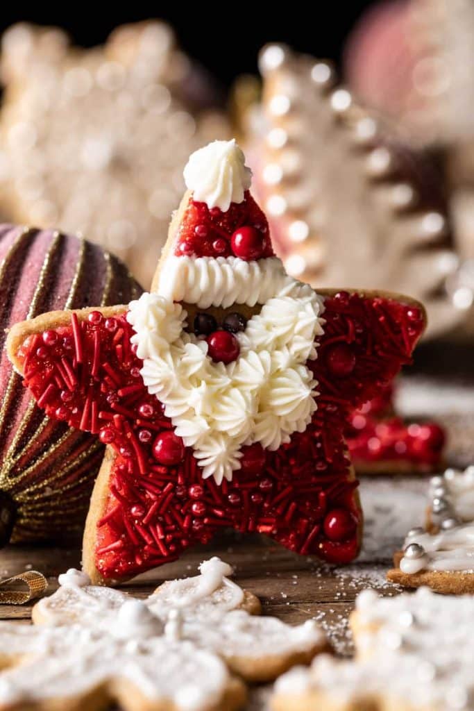 A star-shaped cookie decorated as Santa with red sprinkles and white icing for the beard and hat trim, truly one of the best Christmas cookies. The background reveals a blur of holiday-themed cookies and ornaments, adding to the festive spirit.