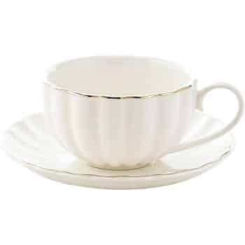 A white ceramic teacup with a scalloped design and gold rim rests on a matching saucer. The cup has a rounded handle, and both pieces have a glossy finish. The setting is simple and elegant.