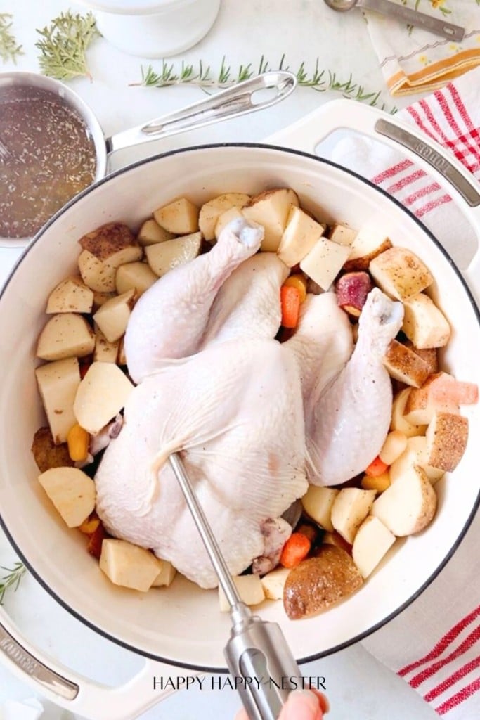 A whole raw chicken, ready for this infused chicken recipe, rests in a white pot surrounded by chopped potatoes and carrots. A meat thermometer is inserted into the bird. Fresh herbs and a dish towel are nearby, while a small pot with liquid brews beside it.