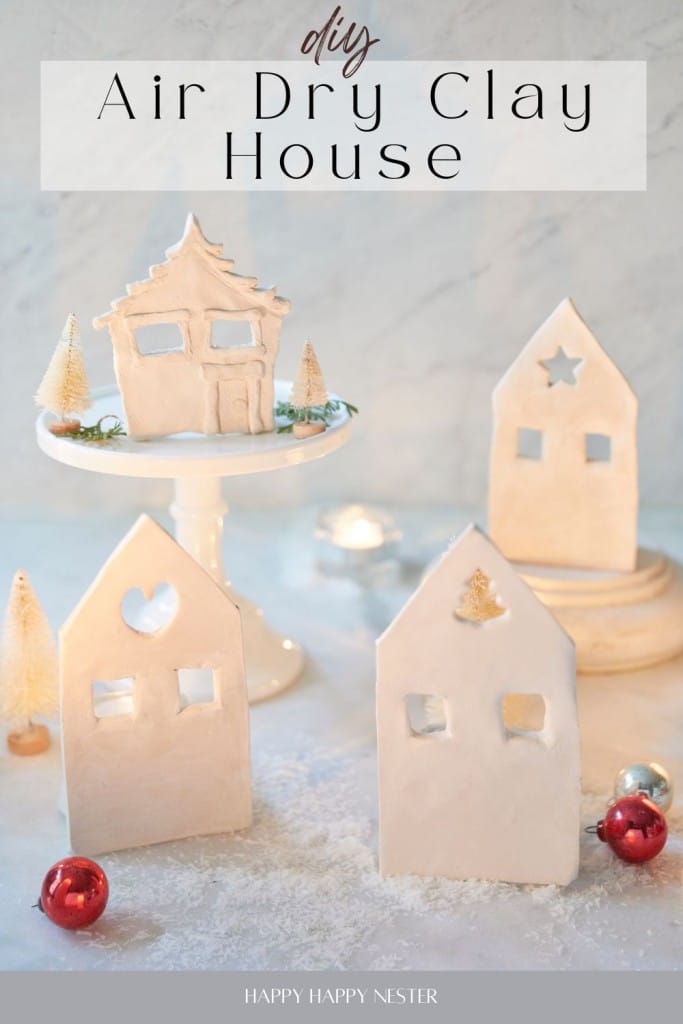 A charming display of handmade air dry clay houses graces a dessert stand and table. Each house, with its uniquely shaped cut-out windows, is surrounded by small decorative trees and holiday ornaments. Text reads "DIY Air Dry Clay House" and "Happy Happy Nester.