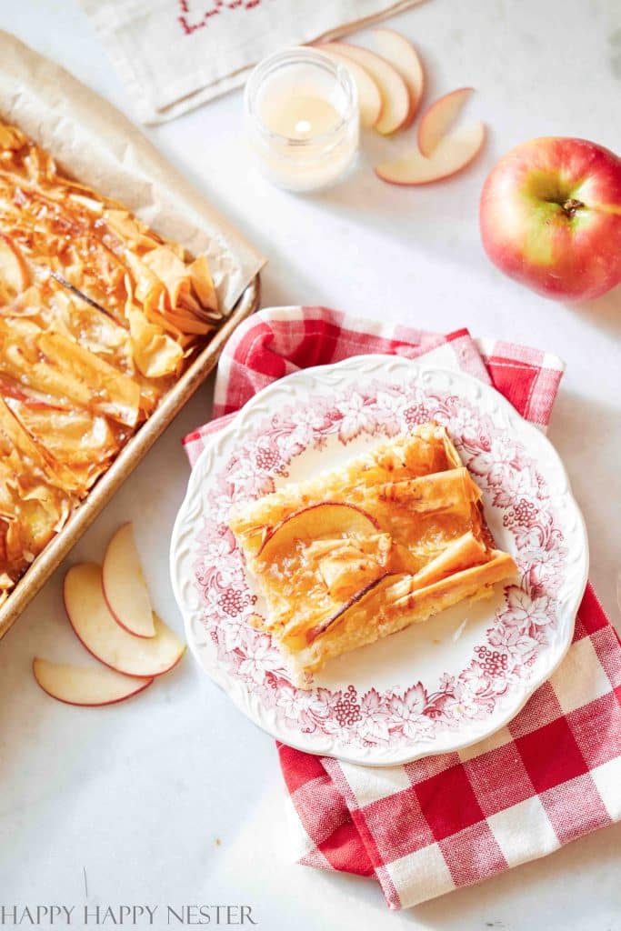 A slice of apple tart, offering cake inspiration, rests on a decorative plate atop a red and white checkered cloth. Nearby, whole and sliced apples accompany the scene. In the background, a baking tray with more tart and a lit candle add to the cozy atmosphere.