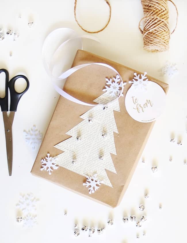 A gift wrapped in brown paper, adorned with a paper Christmas tree and white snowflakes, showcases the art of Christmas crafts for adults. It features a white ribbon and a round "to/from" tag. Scissors, twine, and small bells are scattered on a white surface adding festive charm.