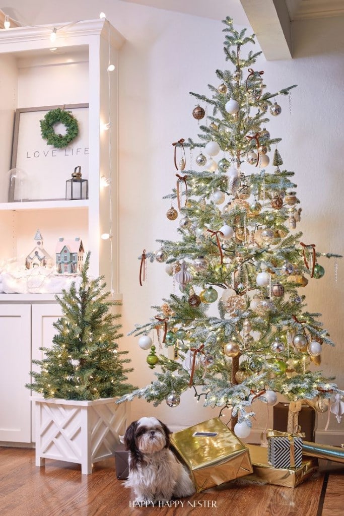 A festive room with two decorated Christmas trees, one large and one small, adorned with white and gold ornaments. A small dog sits beside the trees. Shelves with a wreath and miniature houses are in the background, creating a cozy holiday atmosphere.