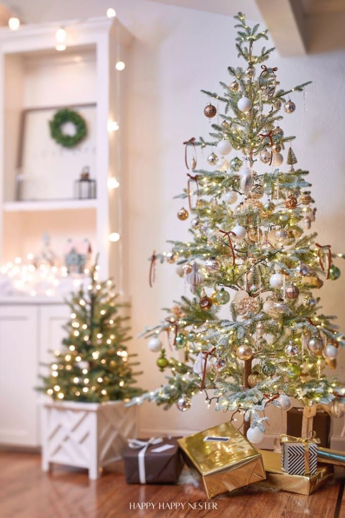 A decorated Christmas tree stands in a cozy room adorned with white and gold ornaments, ribbons, and lights. Nearby, a smaller tree with glowing lights is placed beside wrapped gifts in gold and earth-toned paper. A shelf with holiday decor is in the background.