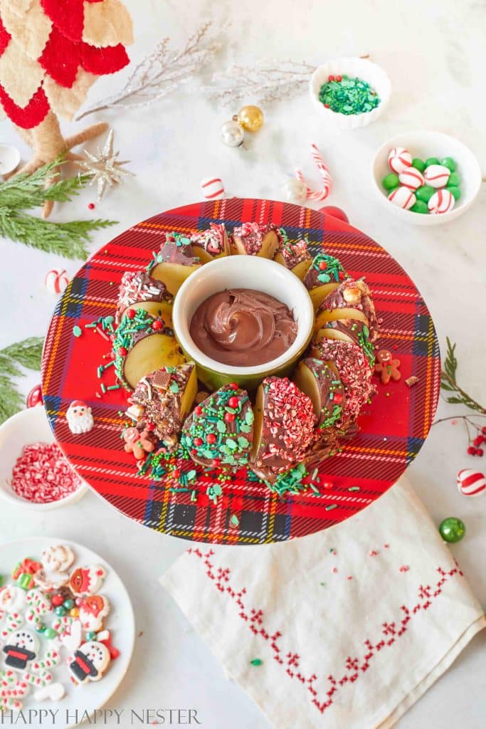 A festive holiday platter features chocolate Christmas treats like apple slices dipped in chocolate and coated with colorful sprinkles, all arranged around a bowl of chocolate sauce on a red plaid plate. Decorated cookies and candies are scattered around the table.