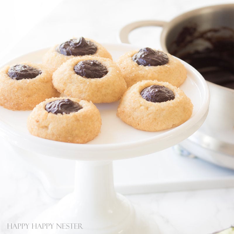 A white pedestal holds six of the best Christmas cookies with chocolate centers, while a pot of extra chocolate tempts in the background on a marble countertop. The image is bright and has a soft, inviting look.