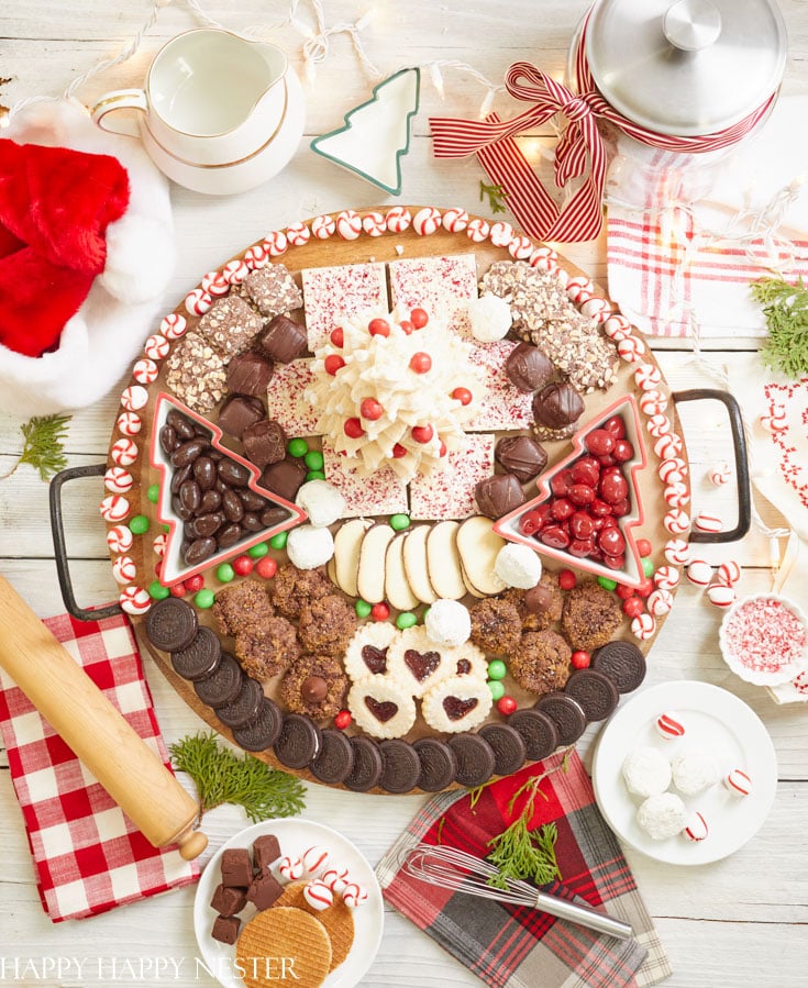 A festive dessert platter featuring an array of chocolate Christmas treats, classic cookies, peppermint candies, and holiday-themed sweets arranged around a centerpiece on a wooden table. Decorated with sparkling ornaments, a rolling pin, and a red-and-white plaid cloth nearby.