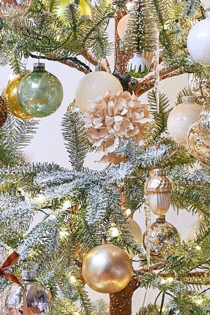 A beautifully decorated Christmas tree with brown and gold ornaments, including silver and green balls, a small Christmas tree ornament, and dried hydrangea. Snow-dusted branches and twinkling lights complete the festive look.