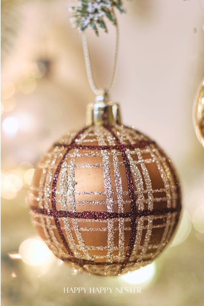 Close-up of a glittery plaid ornament in brown and gold hanging from a branch. The background is softly blurred with warm lights, enhancing the festive mood.