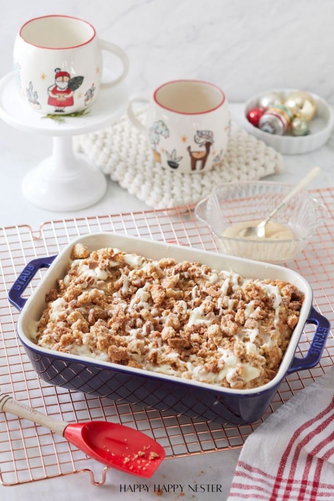 A blue baking dish filled with a crumbly dessert topped with icing sits on a cooling rack. Nearby, two festive mugs with holiday designs are on a stand, along with holiday ornaments and a bowl of glaze. A red spatula rests beside the dish.