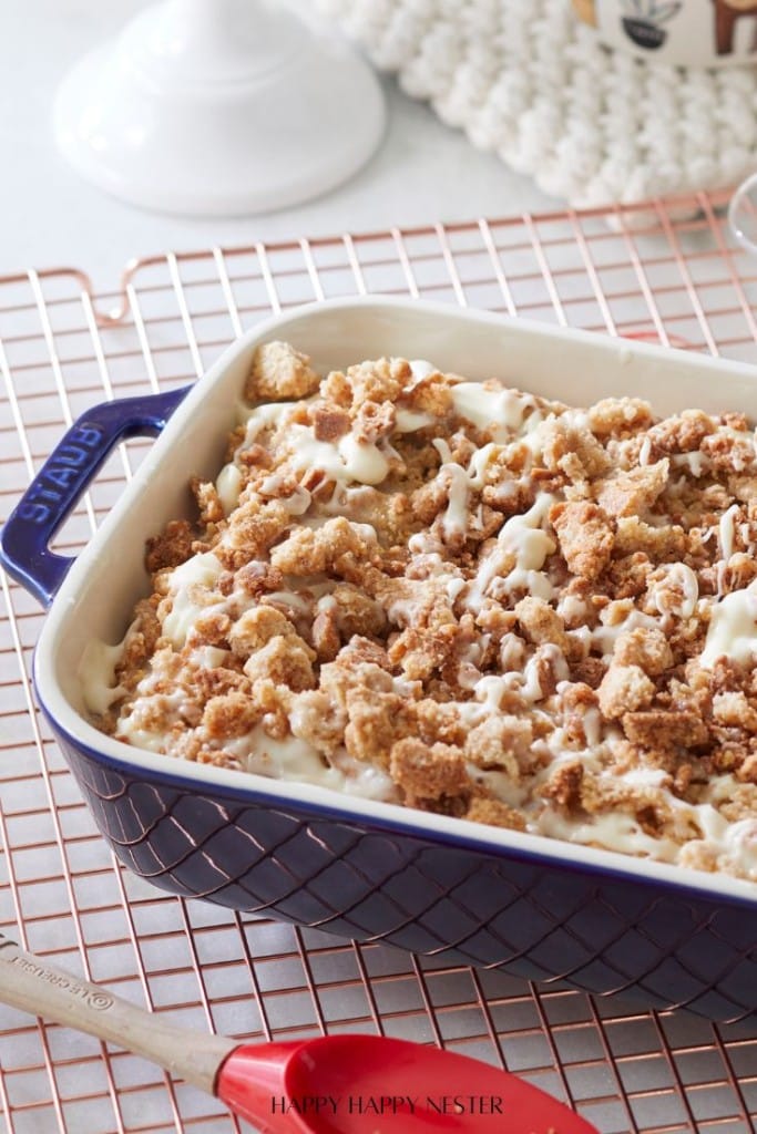 A blue baking dish filled with a delicious dessert, reminiscent of a Coffee Crumb Cake recipe, is topped with crumbled streusel and drizzled with icing. It sits on a wire rack beside a red-handled spatula. A cake stand and textured cloth are artfully blurred in the background.