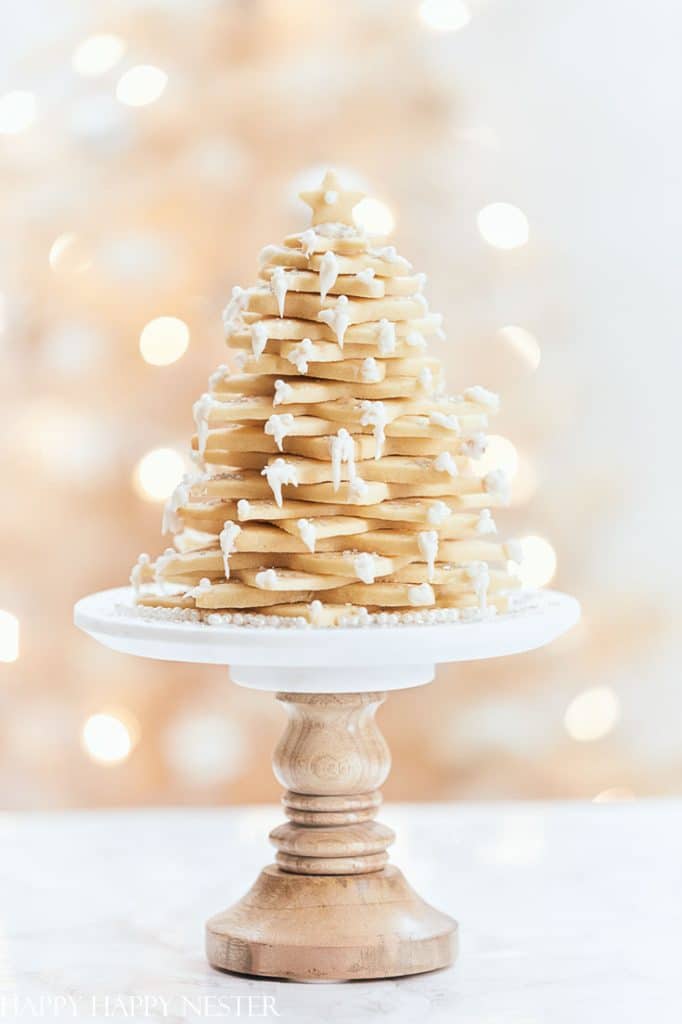 A stack of star-shaped sugar cookies, hailed as the best Christmas cookies, arranged in a tree shape and topped with icing drizzles and powdered sugar. The cookies sit elegantly on a wooden cake stand against a blurred, festive background.