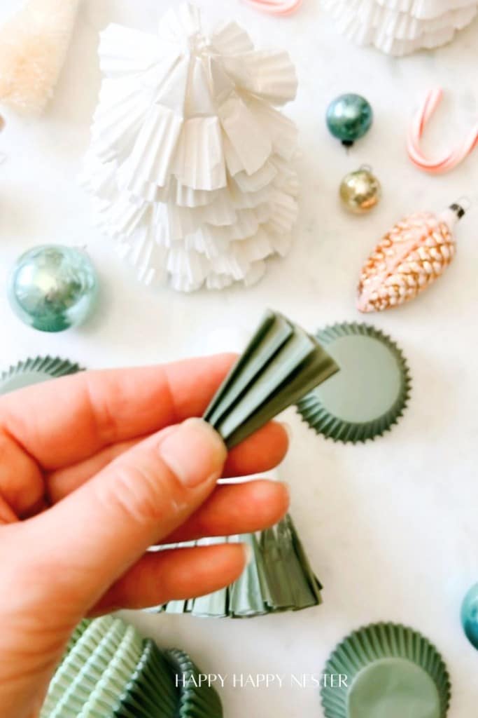 A hand holding a small green paper craft in front of a white textured cone resembling a tree. Surrounding are decorative items, including green ornaments, a pinecone ornament, and candy canes on a marble surface. Text reads "Happy Happy Nester.