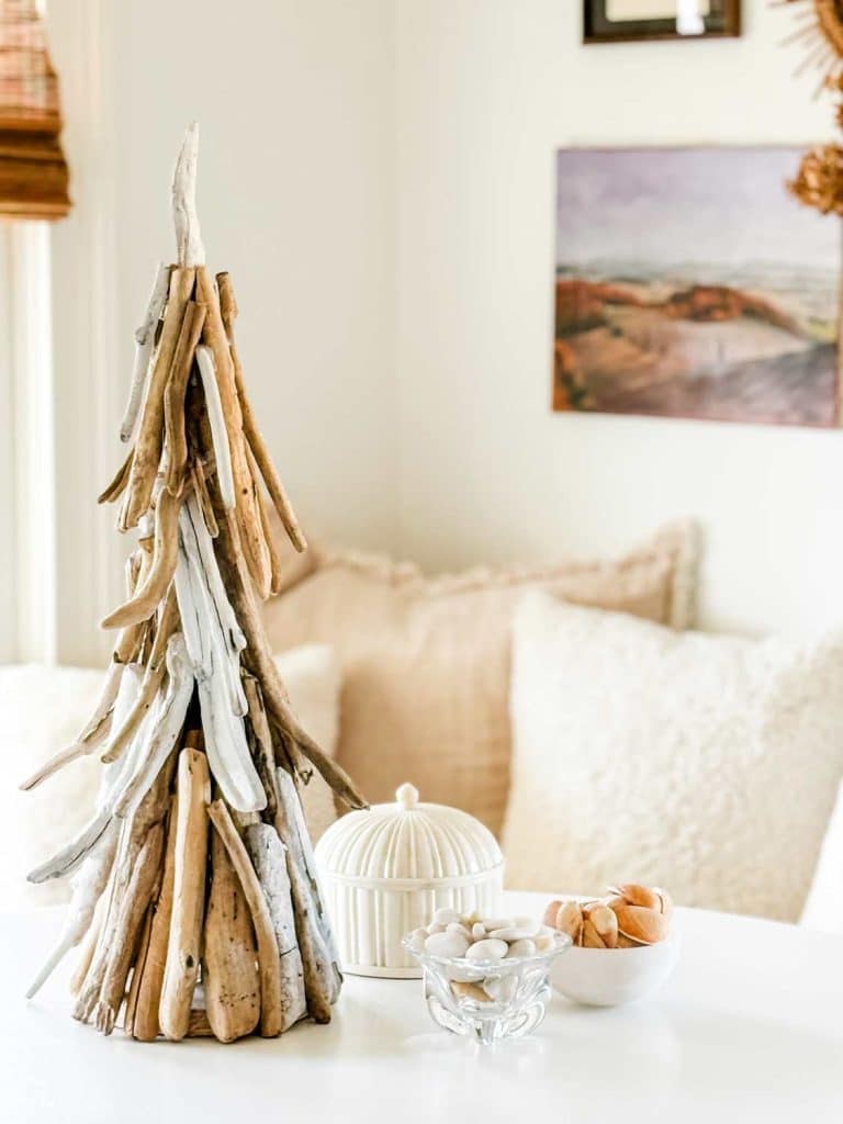 A decorative driftwood tree stands on a white table beside a paper Christmas tree and a white jar with a lid. Two small bowls hold white stones and seashells, while the background showcases beige cushions and a framed landscape photo.