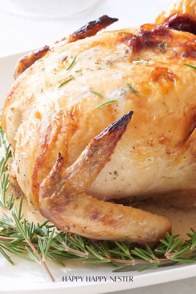 A close-up of a roasted chicken garnished with sprigs of rosemary on a white plate. The chicken is golden brown with crispy skin, seasoned with herbs and pepper, creating an appetizing presentation.