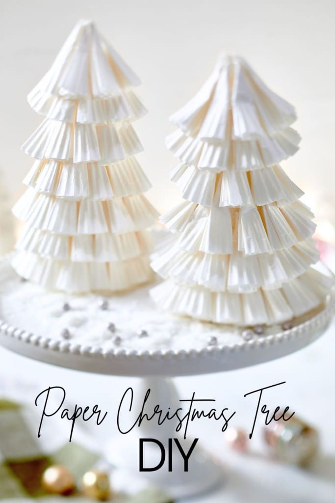 Two paper Christmas trees crafted from white cupcake liners grace a cake stand with faux snow, capturing the essence of a cozy Paper Christmas Tree DIY. Holiday decorations softly blur in the background, adding to the festive allure.