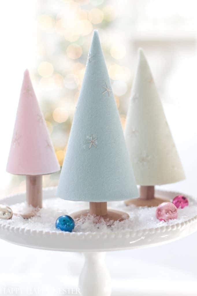 Three pastel-colored cone-shaped decorations resembling Christmas trees are displayed on a white platter with fake snow and small colorful ornaments, offering charming holiday craft ideas. The background has a soft, blurred festive atmosphere.