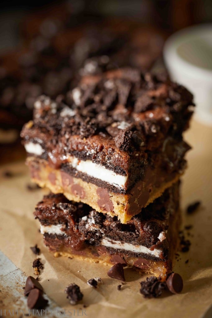 A stack of dessert bars with layers of cookie dough, Oreos, and brownie evokes the essence of irresistible chocolate Christmas treats. Topped with crushed cookies and chocolate chips, their gooey texture and rich chocolate topping shine on parchment paper.