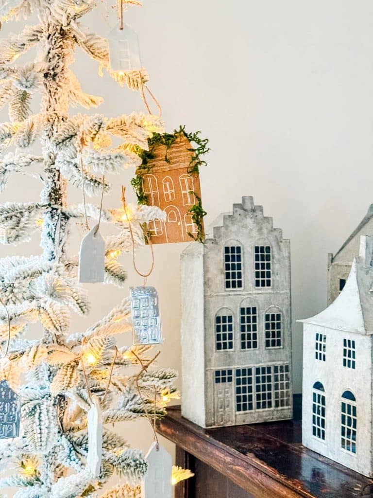 A snowy Christmas tree adorned with small house ornaments and brown and gold decorations stands next to decorative wooden houses on a table. Soft, warm lights illuminate the scene, creating a cozy holiday atmosphere.