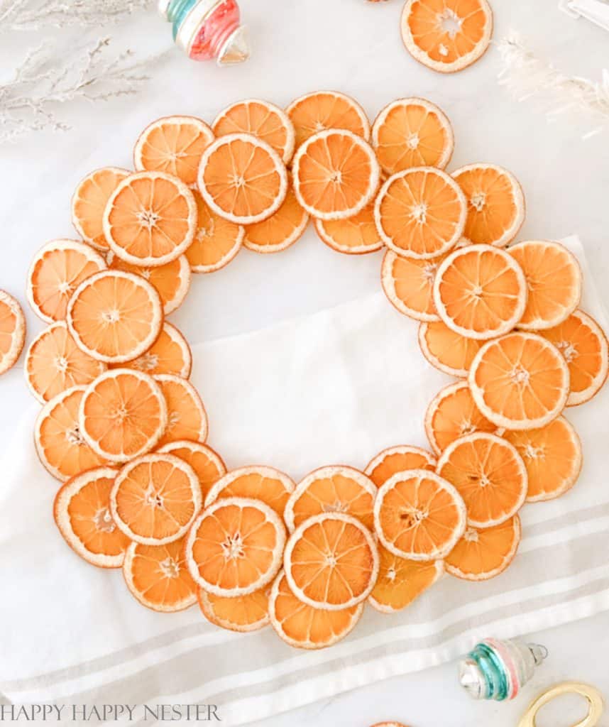 A decorative wreath made of dried orange slices is artfully arranged on a white surface, perfect for Christmas crafts for adults. The orange slices form a circular pattern, with scattered additional slices and a few colorful ornaments adding festive flair.