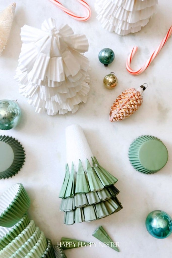 A festive flat lay featuring DIY Christmas trees made from green and white cupcake liners, surrounded by candy canes, shiny ornaments, and a pinecone decoration on a marble surface.