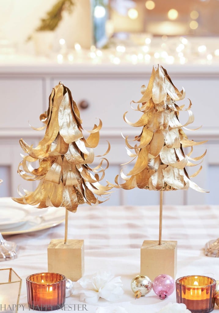 Two gold metallic Christmas tree decorations stand on wooden blocks, showcasing a charming example of Christmas crafts for adults. They're surrounded by candles and ornaments on a festive table. Twinkling lights and a blurred background create a cozy holiday ambiance.