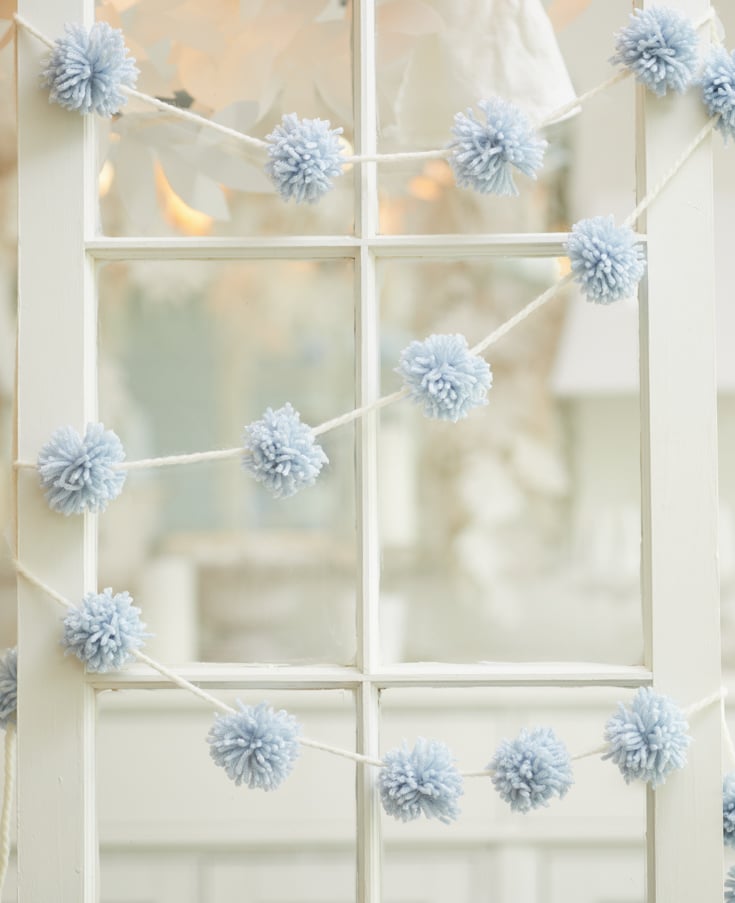 A white window frame is adorned with a garland of light blue yarn pom-poms, perfect for holiday craft ideas. The background is softly blurred, creating a serene and decorative scene.