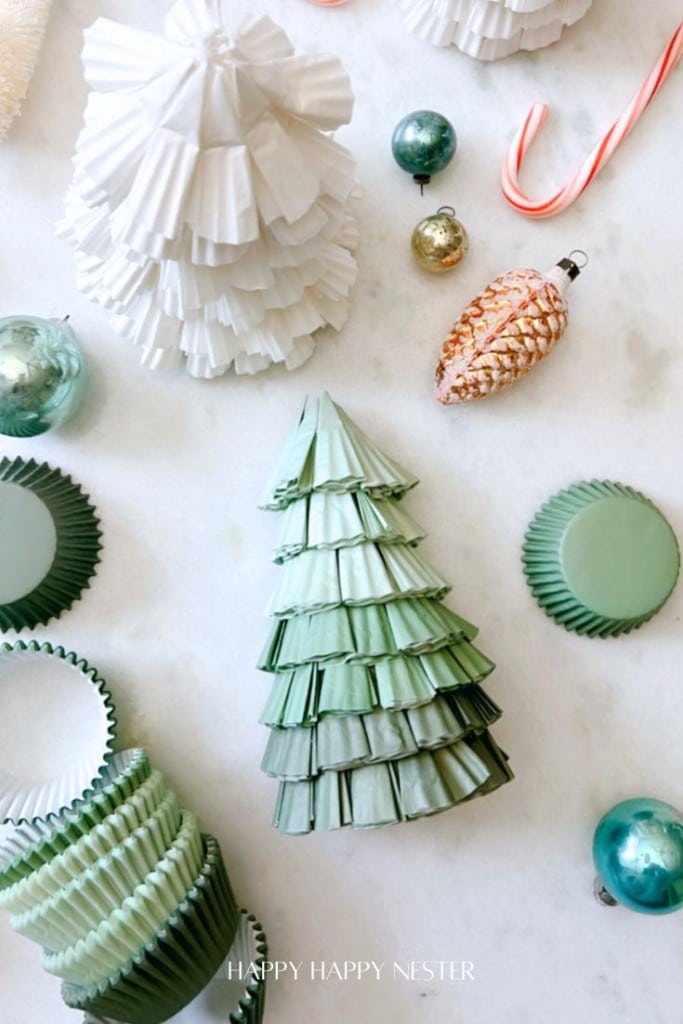 Crafted Christmas trees made from stacked cupcake liners in shades of green and white are surrounded by ornaments and a candy cane on a white surface.