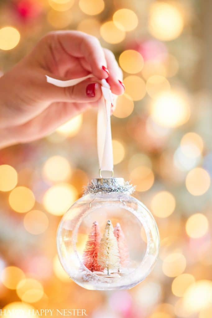 A hand with red nail polish holds a clear Christmas ornament containing small pink and yellow miniature trees, a perfect addition to your holiday craft ideas. The background is blurred with warm golden bokeh lights, creating a magical festive ambiance.