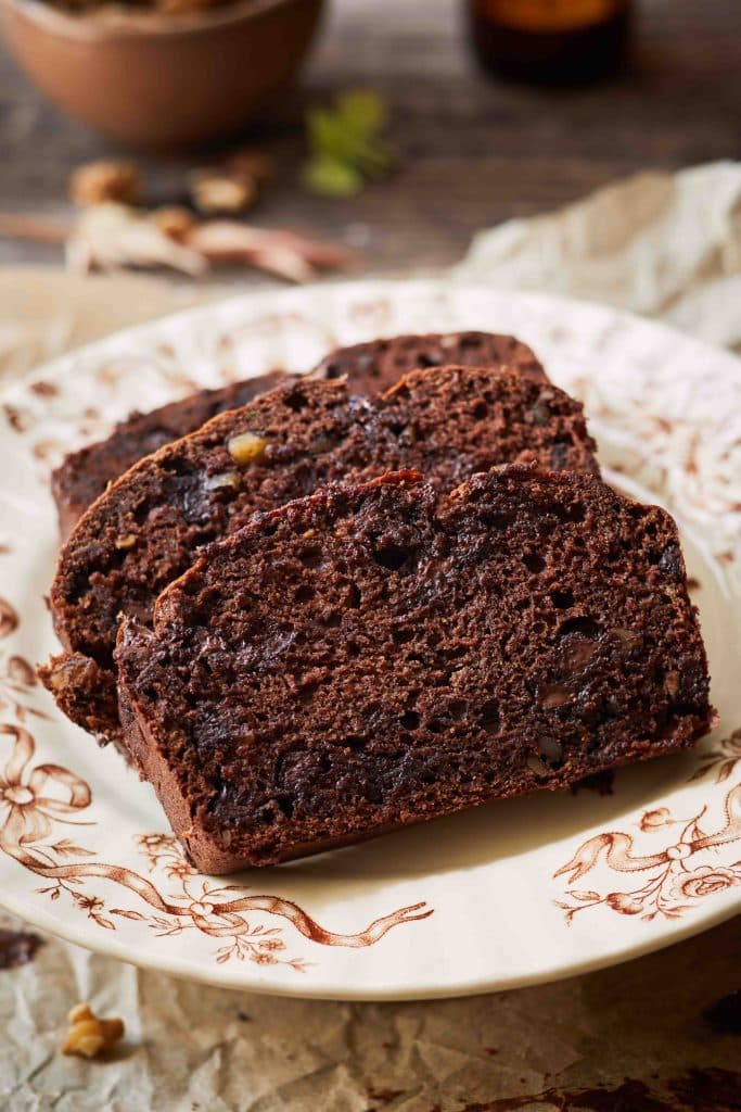 Three slices of chocolate bread, perfect for Christmas treats, rest on a decorative plate atop a rustic surface, with walnuts in the background. The bread appears moist and rich, featuring visible chunks of chocolate and nuts.