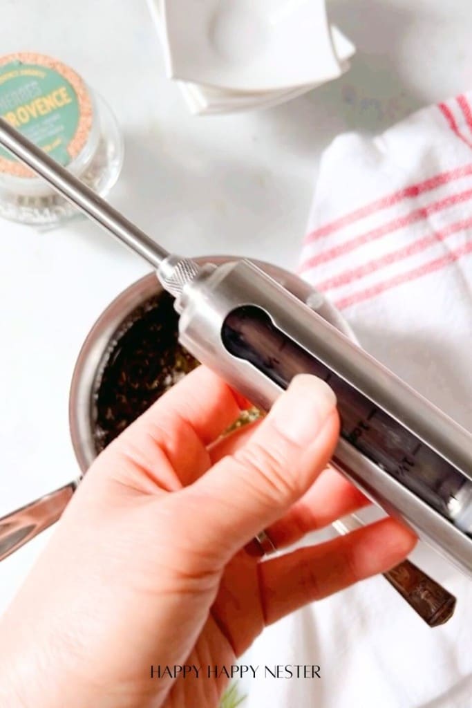 A hand holds an oil sprayer above a bowl filled with herbs, ready for the infused chicken recipe. A red-striped towel and a jar labeled "Herbes de Provence" are visible in the background.