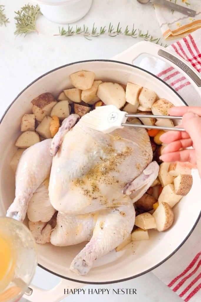 A hand brushes olive oil on an infused chicken recipe nestled in a white pot, surrounded by chopped potatoes and vegetables. A striped dish towel and rosemary sprigs add a touch of charm to the scene.