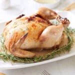 A roasted, infused chicken recipe graces a white platter, garnished with rosemary sprigs. A fork rests beside the platter. In the background, a small plate of butter and a striped napkin add charm to this delectable scene.