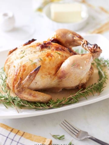 A roasted, infused chicken recipe graces a white platter, garnished with rosemary sprigs. A fork rests beside the platter. In the background, a small plate of butter and a striped napkin add charm to this delectable scene.