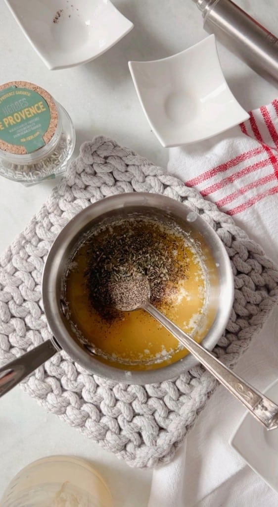 A stainless steel pot with melted butter, herbs, and spices sits on a knitted grey mat, preparing for an infused chicken recipe. A spoon rests in the pot. Nearby, a jar of "Herbs de Provence," a red-striped cloth, and a small white dish add charm to the scene.