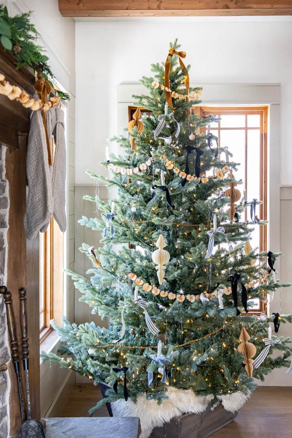 A decorated Christmas tree stands in a cozy room beside a stone fireplace, showcasing brown and gold ribbons alongside white and black ornaments with glowing lights. Stockings hang from the mantel, while a window provides natural light to the festive decorations.