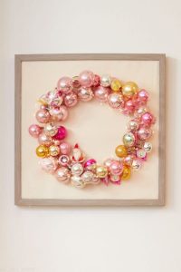 A decorative wreath made of pink, gold, and silver Christmas ornaments is framed and hung on a wall. This delightful piece, perfect for those who love Christmas crafts for adults, features shiny ornaments of varying sizes that create a festive and elegant display.