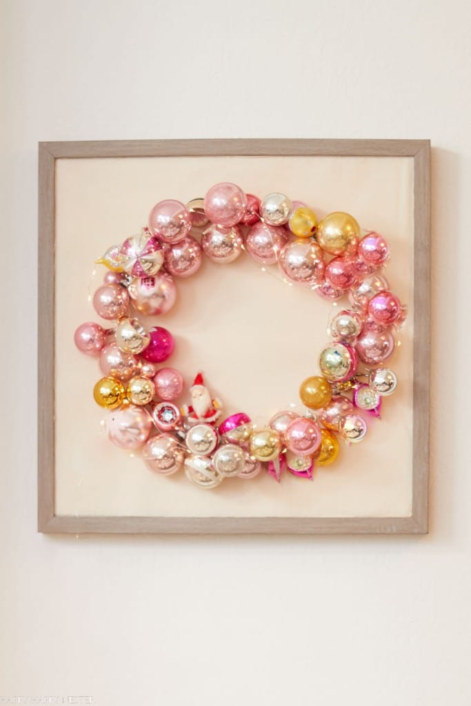 A decorative wreath made of pink, gold, and silver Christmas ornaments is framed and hung on a wall. This delightful piece, perfect for those who love Christmas crafts for adults, features shiny ornaments of varying sizes that create a festive and elegant display.