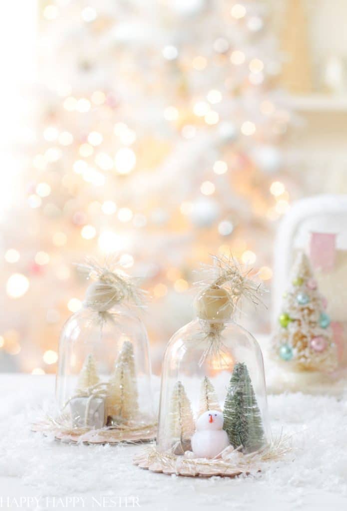 Two small glass domes showcase miniature Christmas scenes with trees and a snowman on a snowy base. In the background, a softly lit Christmas tree and another small decorated tree enhance the warm, festive atmosphere. Perfect inspiration for holiday craft ideas.