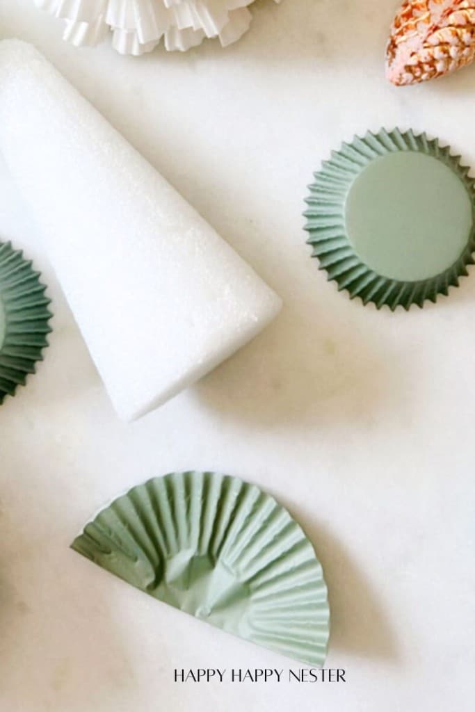 Various crafting materials, including a white foam cone, green cupcake liners, and a pine cone, are arranged on a light surface with the text "Happy Happy Nester" at the bottom.