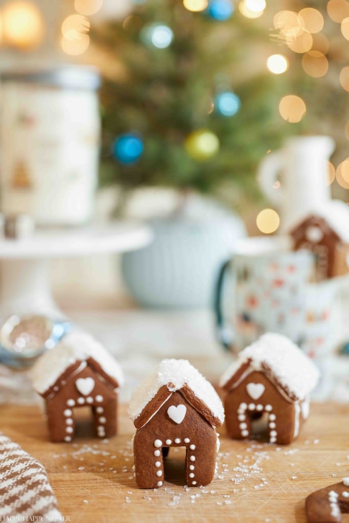 Three decorated gingerbread houses, hailed as the best Christmas cookies, stand proudly in the foreground with intricate icing details. In the background, a blurred Christmas tree twinkles with lights and ornaments, creating a cozy holiday setting alongside a mug and festive decorations.