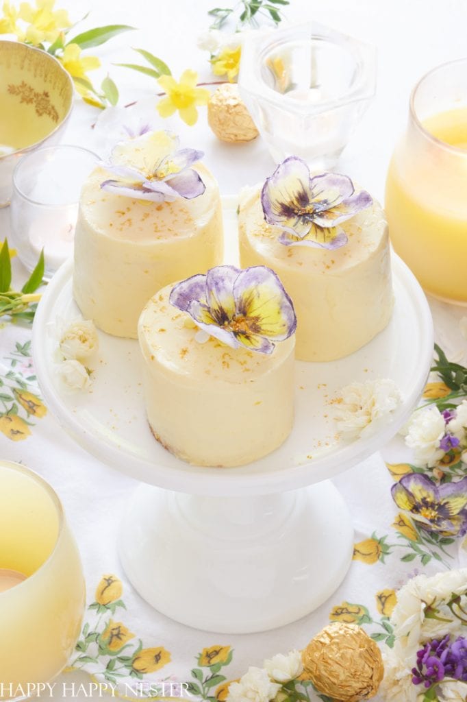 Three small, round cheesecakes topped with purple and yellow edible flowers sit on a white cake stand, offering delightful cake inspiration. The background features yellow floral decorations, candy, and glasses with a light drink.