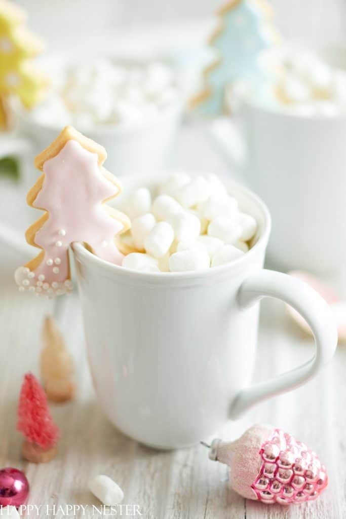 A white mug filled with small marshmallows sits on a table, with a pink tree-shaped cookie, one of the best Christmas cookies, perched on the rim. In the background are holiday decorations, including colorful miniature trees and ornaments.