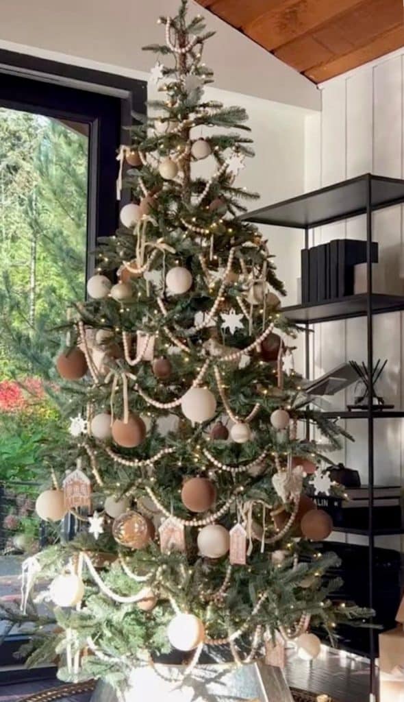 A decorated Christmas tree stands in a modern living room, adorned with brown and gold Christmas tree decorations, wooden stars, and beaded garlands. Sunlight filters through large windows, highlighting the tree's festive charm and the greenery outside.