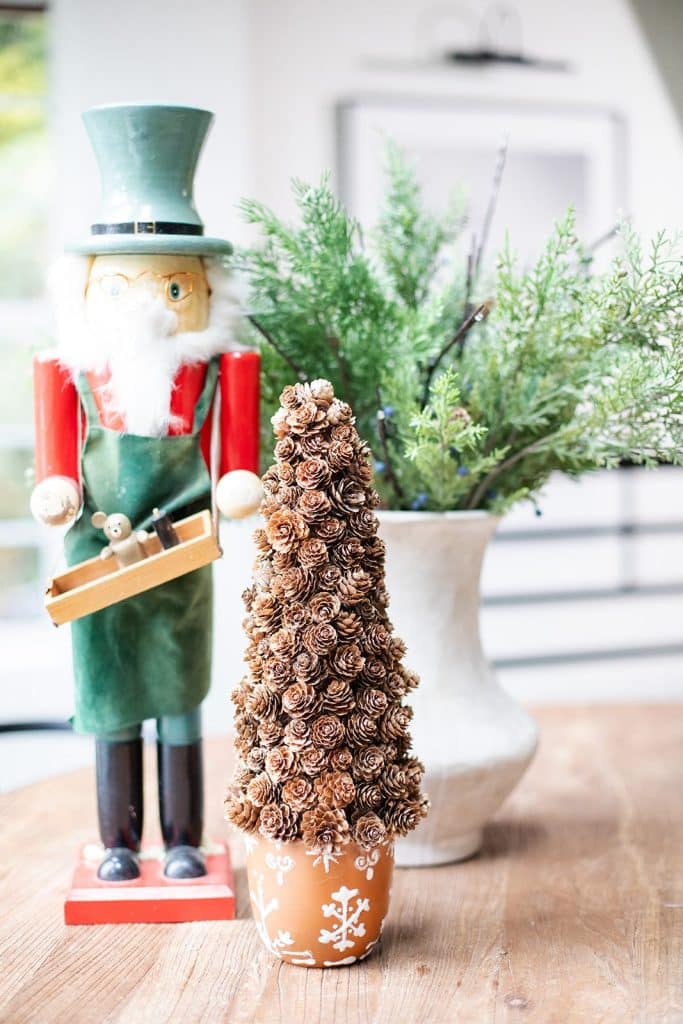 A wooden nutcracker figurine in a green hat and apron stands next to a small pinecone tree in a brown pot with white snowflake designs. Beside it, a charming paper Christmas tree adds holiday cheer, while a vase with greenery graces the background, all set on a rustic wooden table.