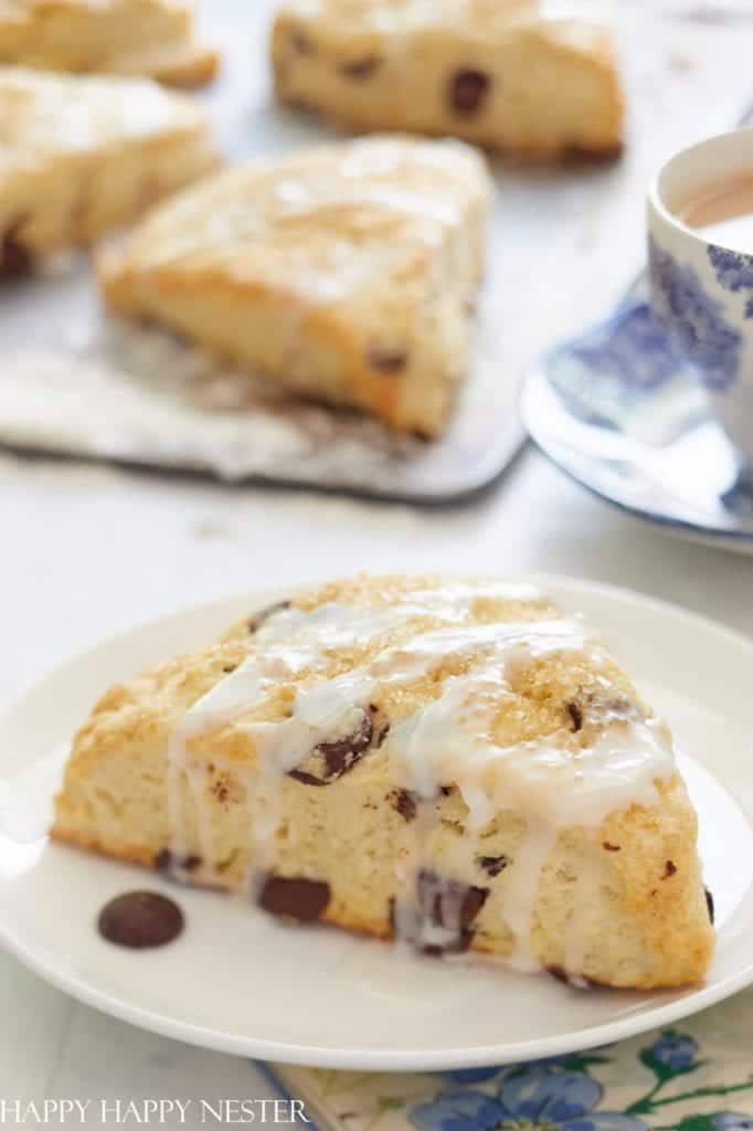 A close-up of a chocolate chip scone drizzled with icing on a white plate. In the background, more festive Christmas treats rest on parchment paper. A floral-patterned teacup is partially visible to the side.