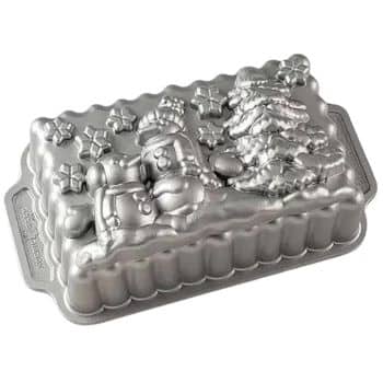 A silver cake pan offers cake inspiration with its embossed designs of a snowman, a decorated Christmas tree, and scattered snowflakes. The rectangular shape features ridged sides and convenient handles on each end.