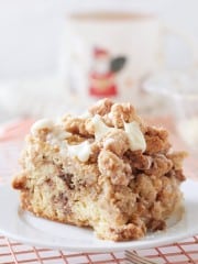 A close-up of a slice of crumbly coffee cake on a white plate showcases the perfect Coffee Crumb Cake recipe. The cake boasts a dense texture with crumbled topping and drizzled icing, while a blurred mug with a festive design adds charm in the background.